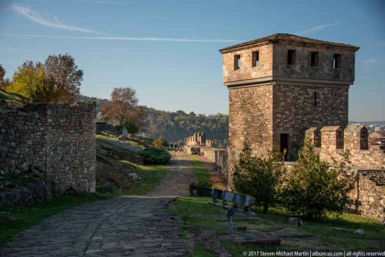 Cobbled fortress lane beside a tall square stone tower and crenellated defensive walls.