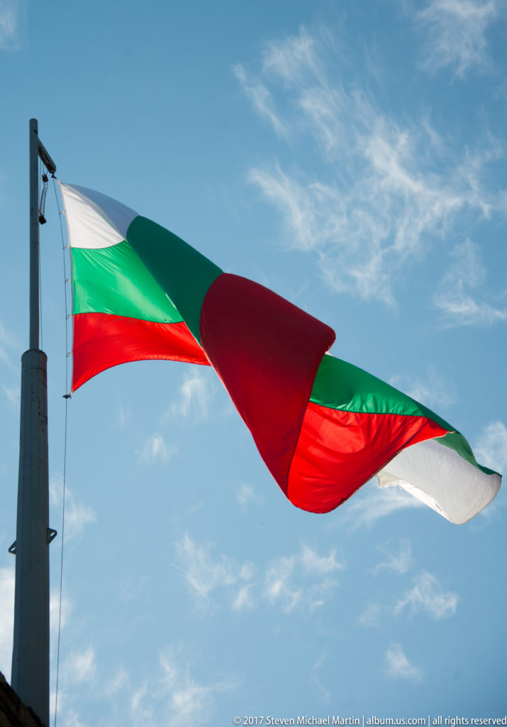 Bulgarian white, green, and red national flag billowing from a tall pole against blue sky.