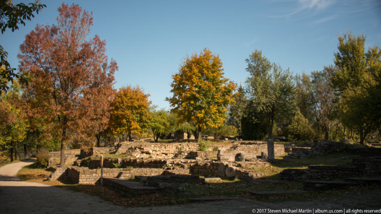 Low stone building foundations spread beneath autumn trees beside paths on the fortress hill.