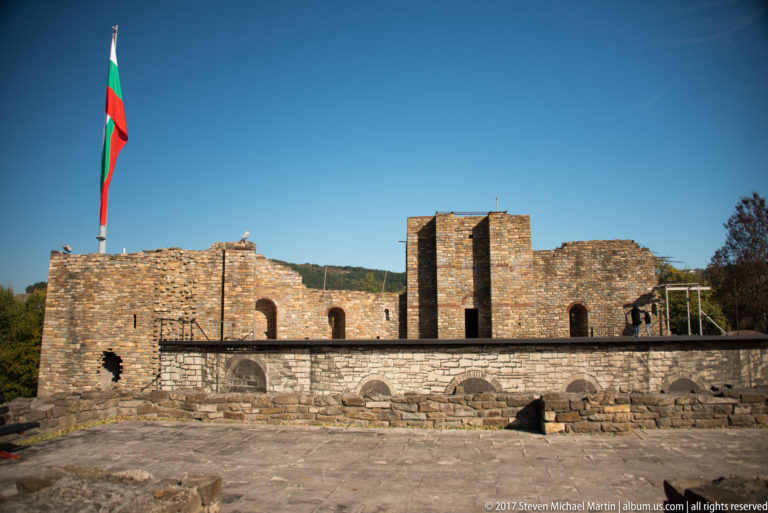 Bulgarian flag above a broad stone ruin with arched openings, tall piers, and carved fragments in front.