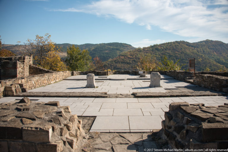Large pale stone-paved terrace with short stone blocks, surrounded by rough fortress foundations and wooded hills.