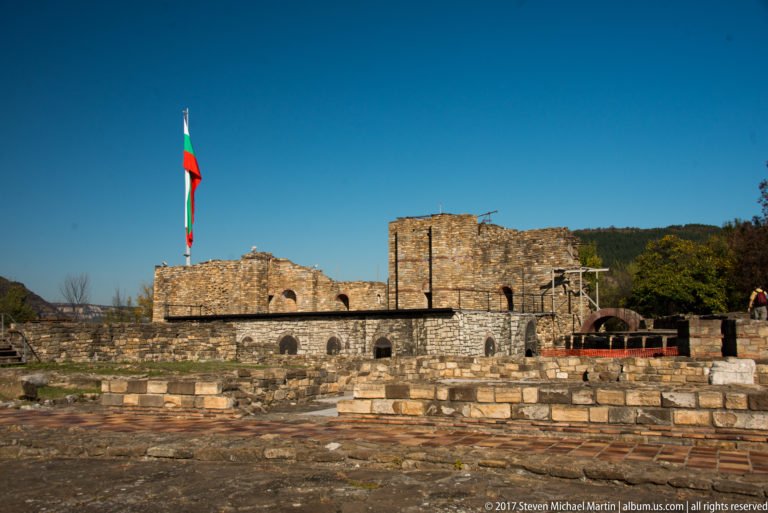 Wide view of stone fortress ruins and foundations beneath a tall Bulgarian flag.