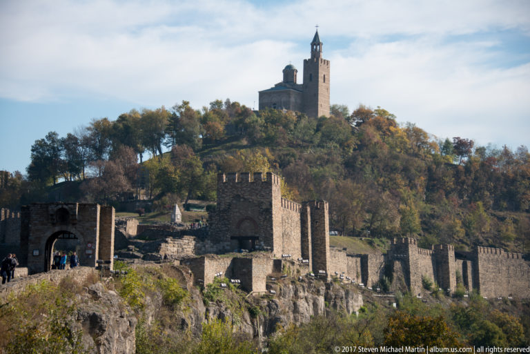 Stone fortress walls and gate towers on a rocky ridge below a church-like hilltop tower.