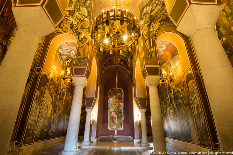 Symmetrical Patriarchal Church interior with pale columns, pointed arches, chandelier, and monumental murals by Teofan Sokerov.
