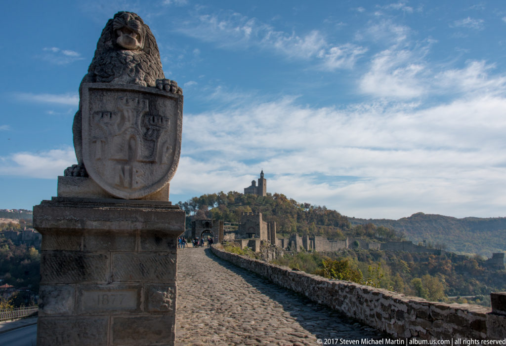 Heraldic stone monument beside a cobbled fortress road leading toward ruins and a hilltop tower.