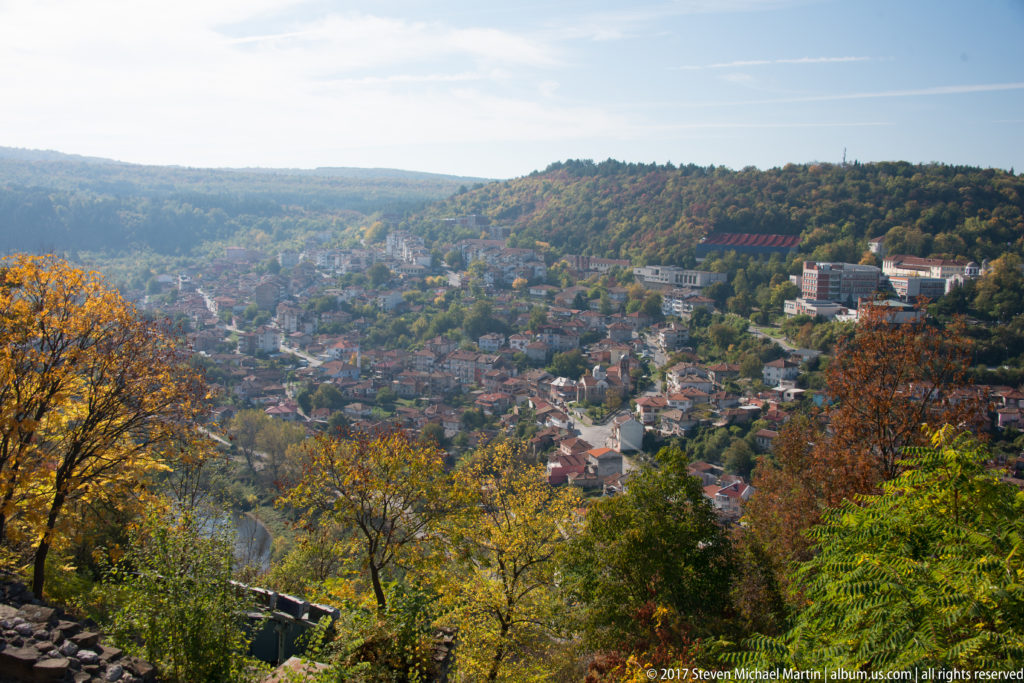 Autumn panorama across Veliko Tarnovo, with red-roofed neighborhoods filling a steep valley beneath wooded hills.