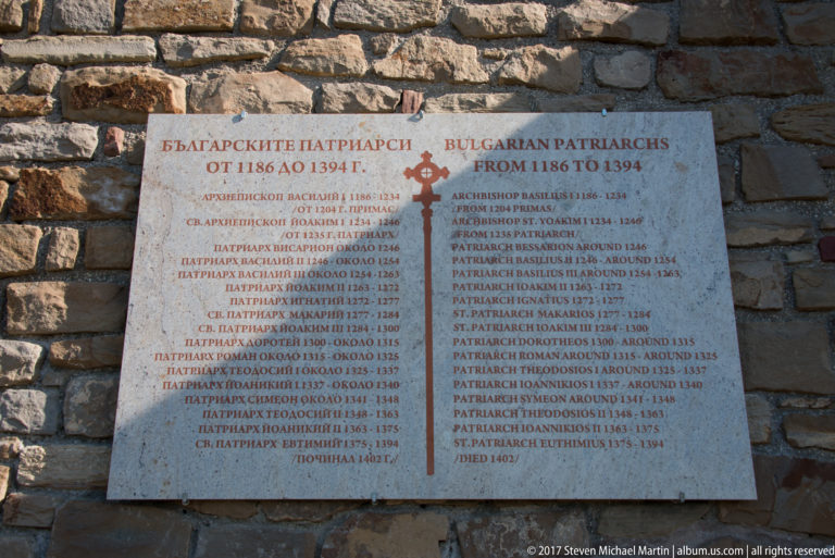 Bilingual stone plaque listing Bulgarian Patriarchs from 1186 to 1394, mounted on rough masonry with a red cross at center.