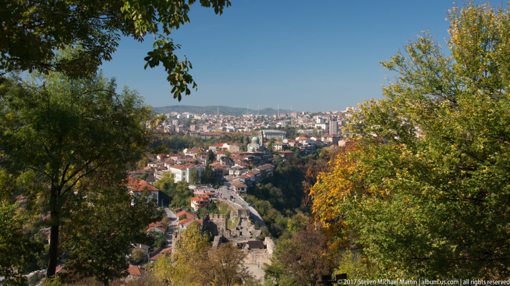 Elevated city view framed by autumn trees, with a descending road and stone walls leading toward dense hillside neighborhoods.