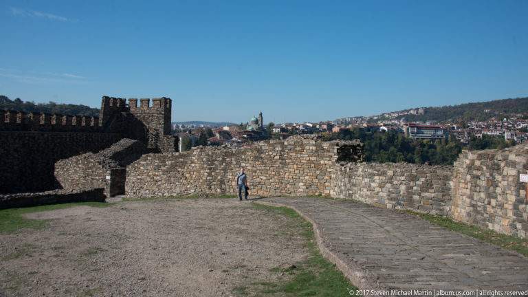 Stone fortress walls and crenellated tower along a curving path on Tsarevets, with one visitor and Veliko Tarnovo beyond.