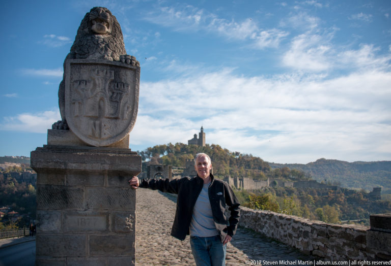 Man standing beside a heraldic stone monument with fortress walls and a distant tower behind him.