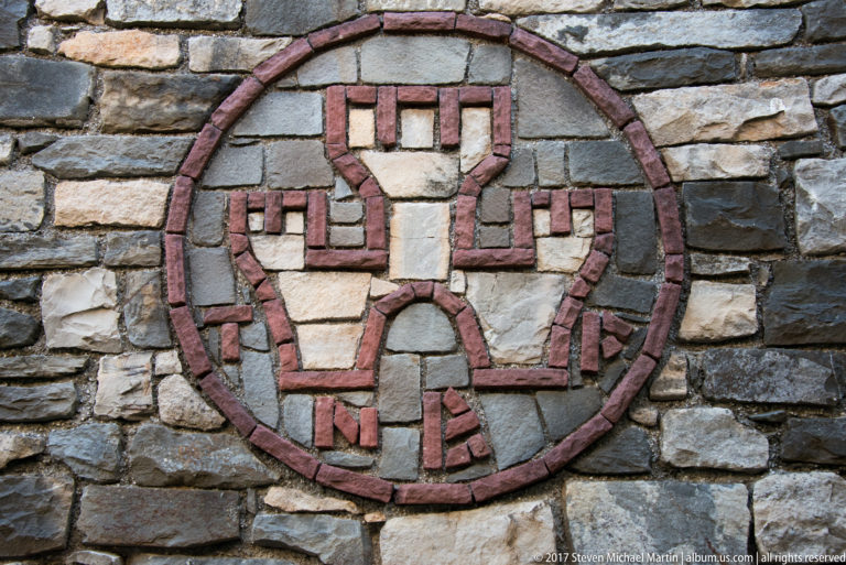 Circular red-brick emblem with a stylized geometric figure set into an irregular gray-stone wall.