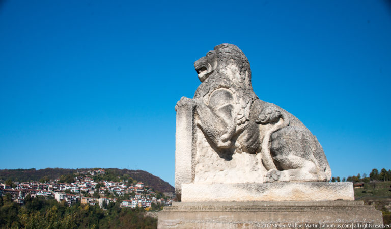 Seated stone lion sculpture on a pedestal in three-quarter profile, with Veliko Tarnovo hillside buildings in the background.