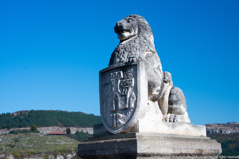 Seated stone lion holding a carved heraldic shield, viewed from a low angle against blue sky and wooded hills.