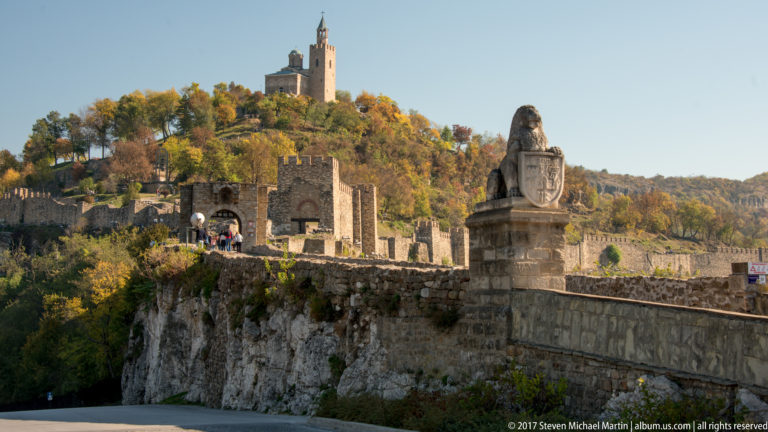Tsarevets approach with stone lion monument, arched fortress gateway and walls, and the Patriarchal Cathedral on the wooded summit.