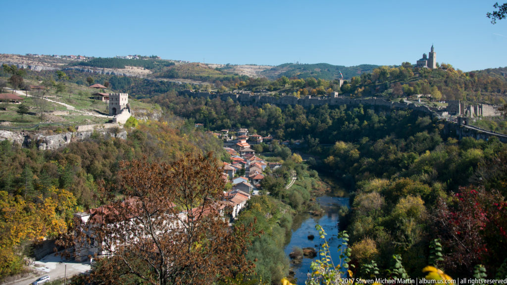 Elevated view over a narrow river winding through a wooded valley, with hillside neighborhoods and a church-topped ridge in the distance.