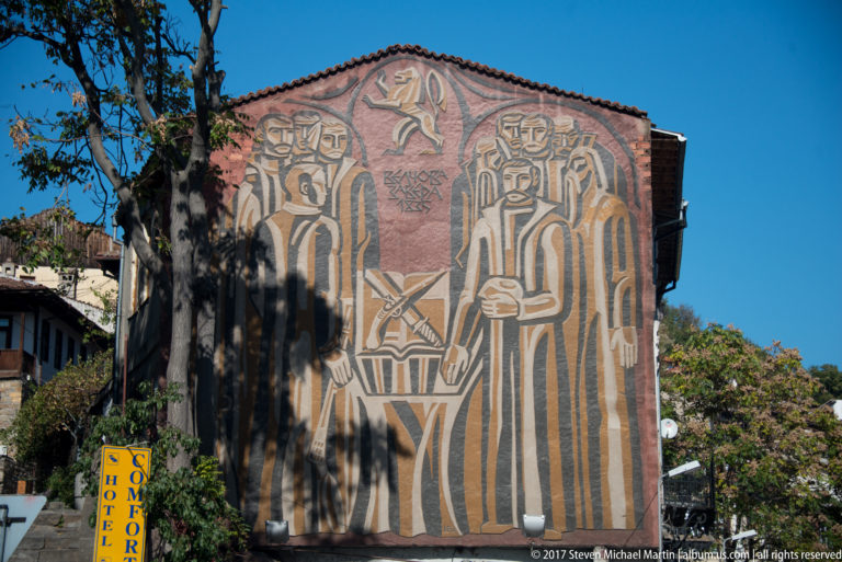 Large exterior mural of elongated robed figures and a central vertical motif covering a building façade under blue sky.