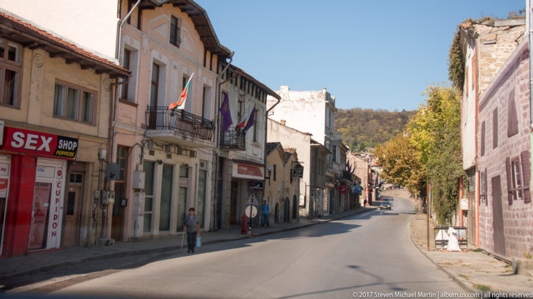 Curving Veliko Tarnovo street lined with older buildings, balconies, shops, parked cars, pedestrians, and Bulgarian flags.