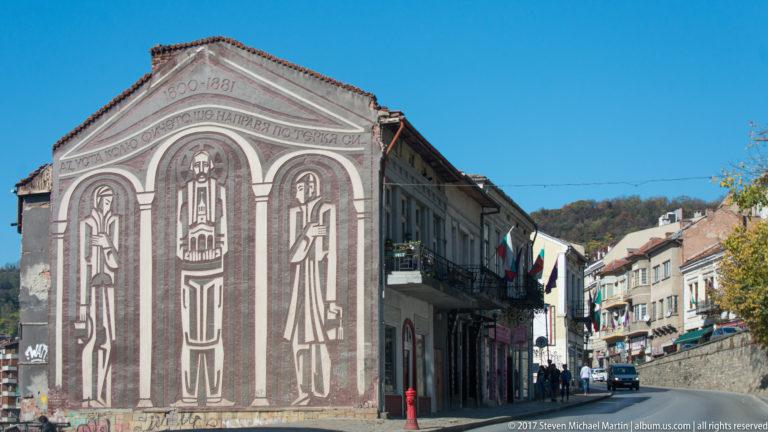 Large symmetrical façade mural with three stylized robed figures, arched motifs, and Cyrillic text above a Veliko Tarnovo street.
