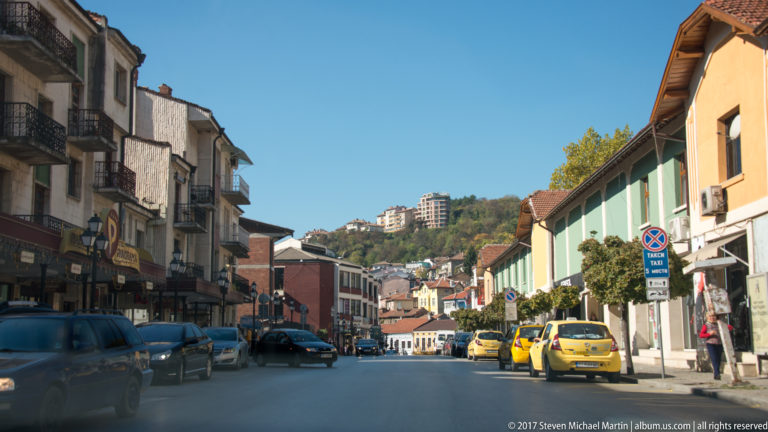 Broad Veliko Tarnovo street with parked cars, yellow taxis, balconies, shops, tiled roofs, and apartment blocks on a wooded slope.
