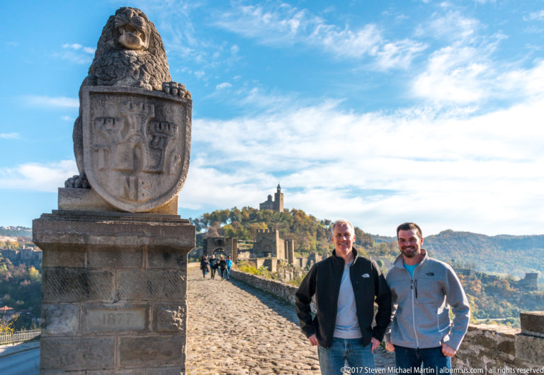 Two visitors beside a tall stone lion-and-shield monument on a cobbled fortress road.