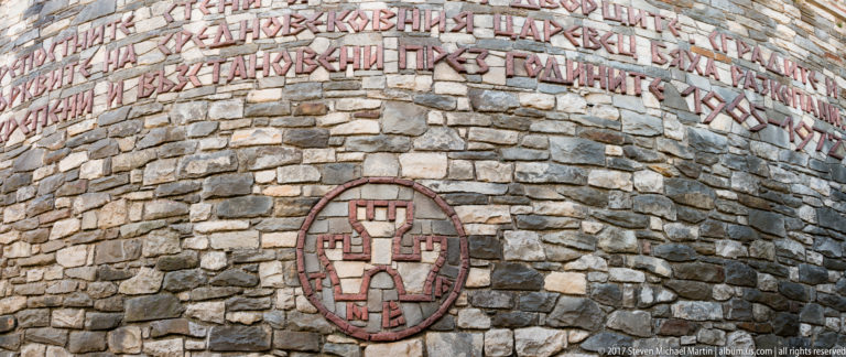 Rough stone wall with raised red Cyrillic lettering above a circular red-brick geometric emblem.