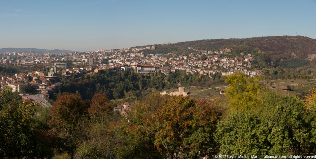 Wide view of Veliko Tarnovo neighborhoods across wooded hills and ravines, framed by autumn trees in the foreground.