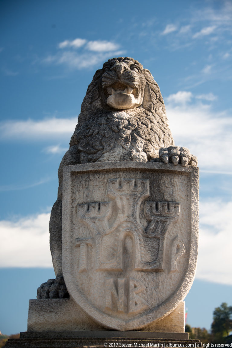 Weathered stone lion crouched above a carved heraldic shield on a masonry monument against blue sky.