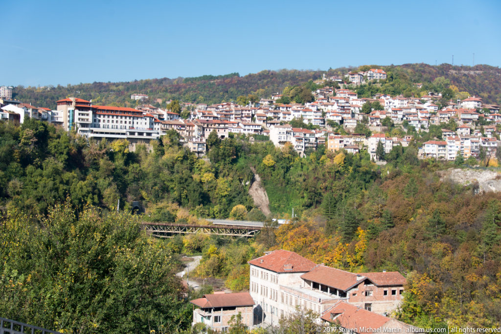 Wide view of Veliko Tarnovo neighborhoods across wooded autumn hills, with larger buildings above a ravine.