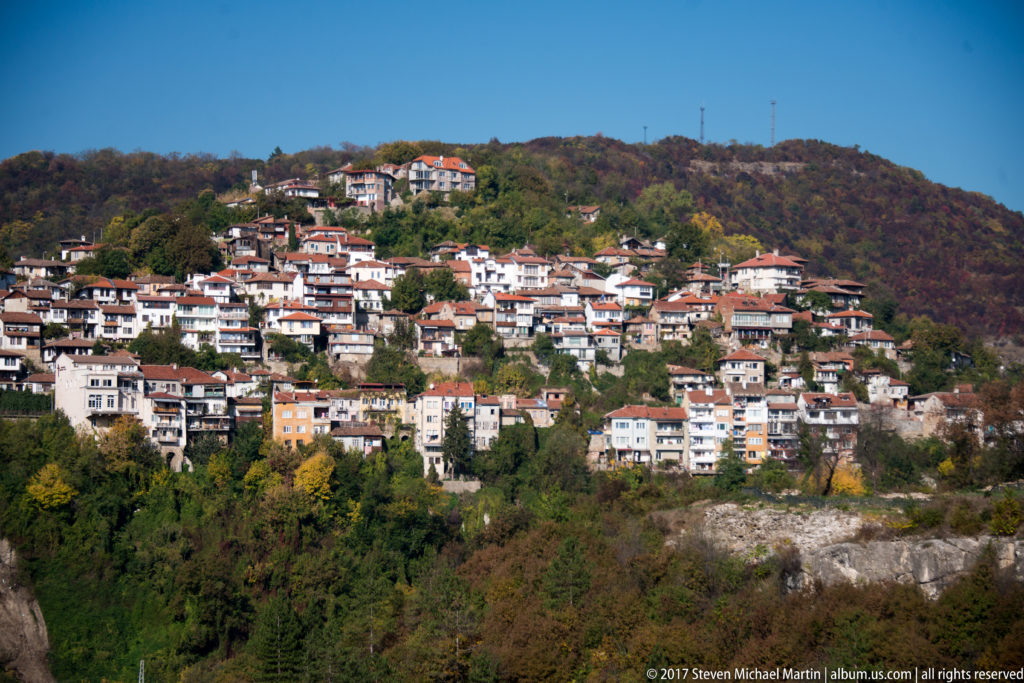 Dense hillside neighborhood of red-roofed houses and apartments surrounded by autumn woodland and exposed rock.