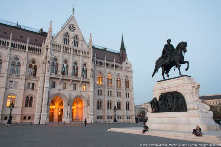 Bronze equestrian monument on a stone pedestal before the Hungarian Parliament’s illuminated façade at blue hour.