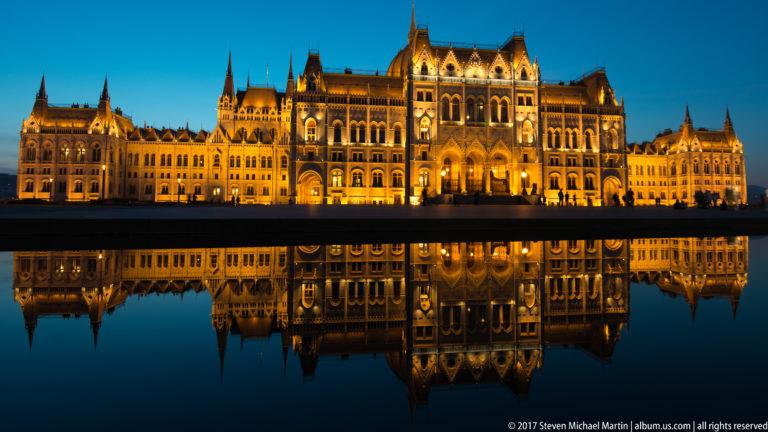 Wide illuminated Hungarian Parliament façade mirrored almost completely in a dark reflecting pool.