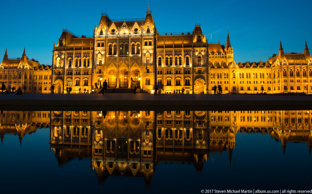 Floodlit Hungarian Parliament with silhouetted visitors and a crisp reflection in the pool below.