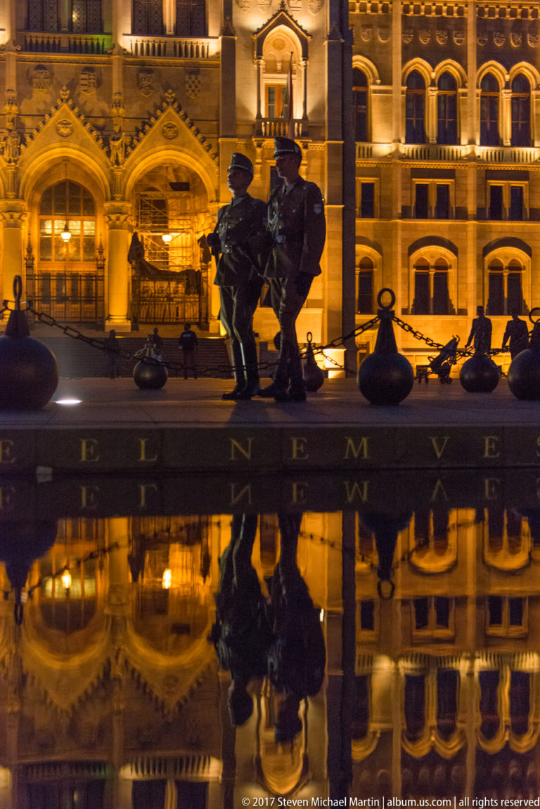 Two uniformed guards beside a dark memorial, with illuminated architecture and their reflections in a shallow pool.