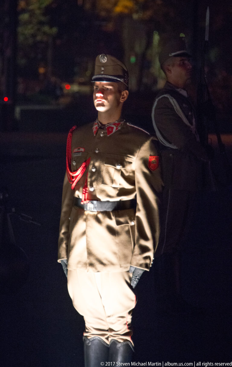 Uniformed ceremonial guard in a peaked cap and red cord standing under a spotlight at night.