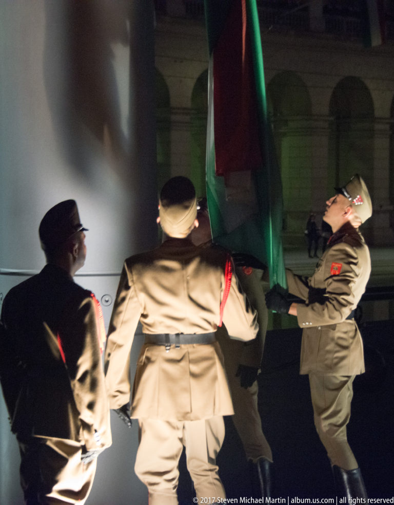 Uniformed guards handling a large red, white, and green Hungarian flag during a nighttime ceremony.