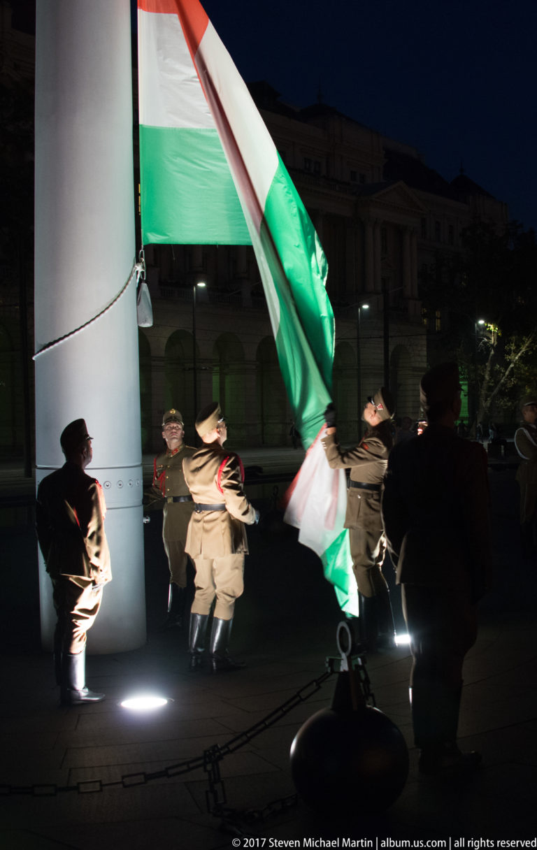 Several uniformed guards position a large Hungarian tricolor beside a tall flagpole at night.