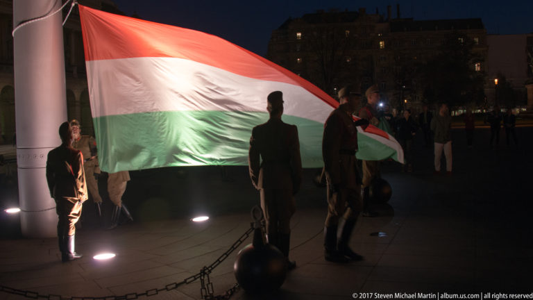 Large Hungarian tricolor spread beside a flagpole as ceremonial guards hold its edges at night.