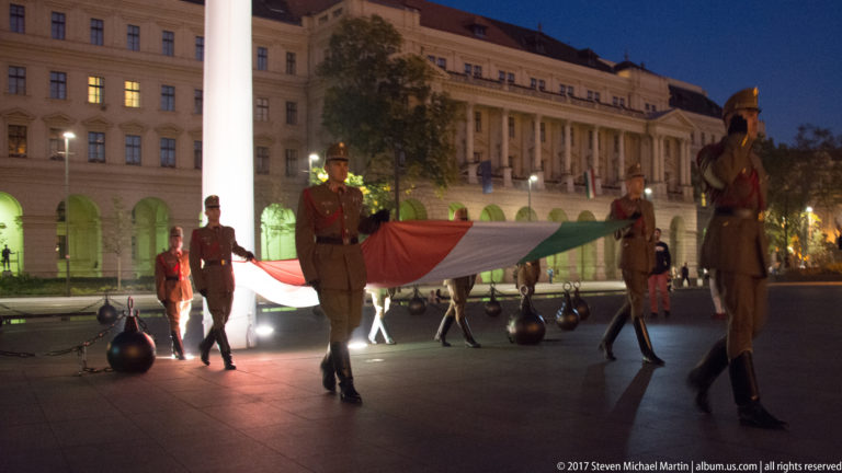 Uniformed guards carrying a large Hungarian flag horizontally across Kossuth Lajos Square at night.