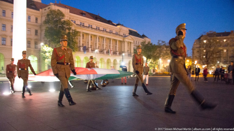 Marching ceremonial guards carry a broad Hungarian tricolor across an illuminated Budapest square at night.