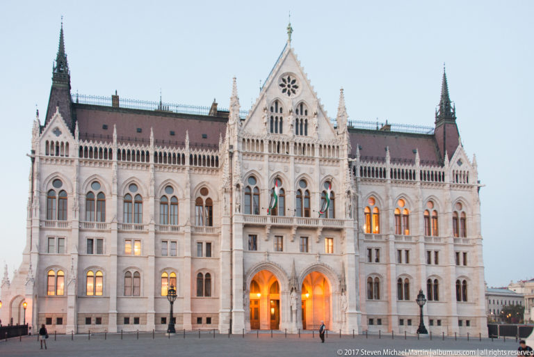 Frontal blue-hour view of the Hungarian Parliament’s ornate central façade with glowing arched entrances.