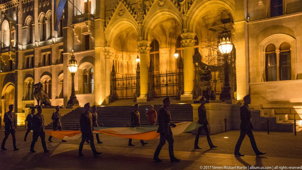 Guards carry a large Hungarian flag past the illuminated arched entrance of the Hungarian Parliament.