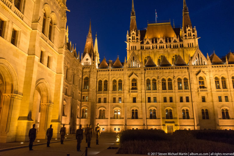 Illuminated side elevation of the Hungarian Parliament with steep gables, pinnacles, and small figures below.