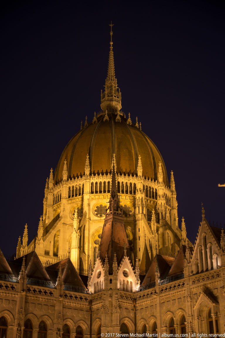 Close night view of the Hungarian Parliament’s illuminated ribbed dome above Gothic Revival roofs and pinnacles.