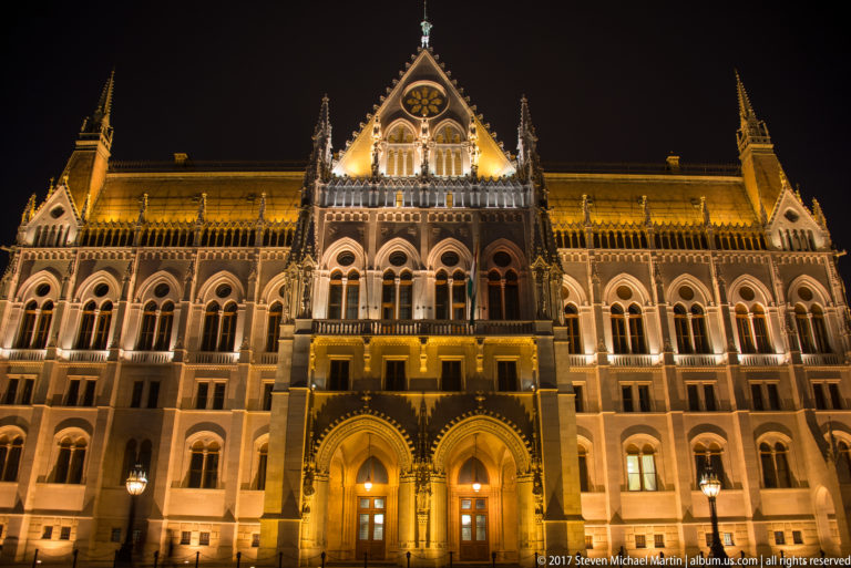 Low-angle night view of an illuminated Hungarian Parliament pavilion with twin arches and a steep central gable.