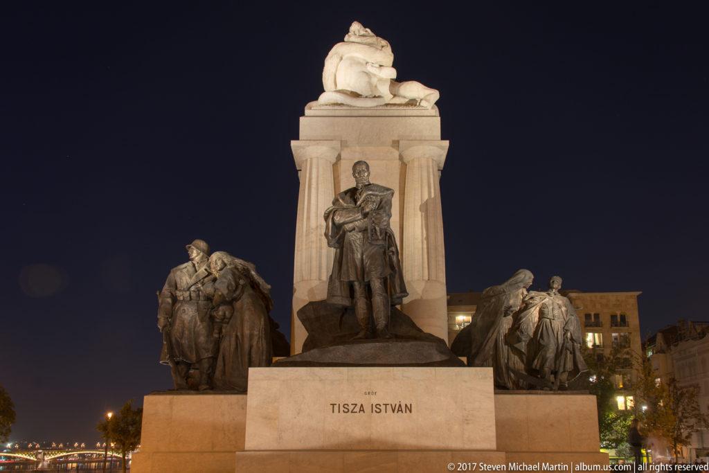 Night view of the István Tisza monument, with a central bronze figure and flanking sculptural groups.