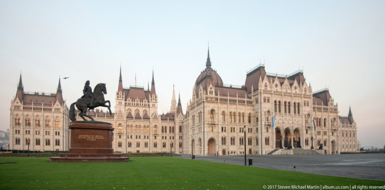 Bronze equestrian monument on a lawn before the broad Gothic Revival façade of the Hungarian Parliament.