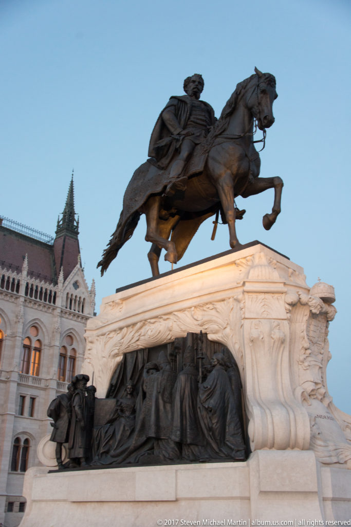 Bronze horse-and-rider monument on a pale pedestal with sculptural reliefs beside the Hungarian Parliament.