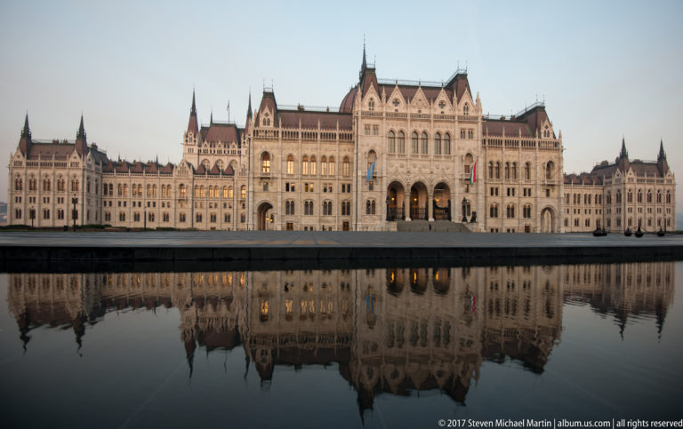 Wide daylight view of the Hungarian Parliament and its pale façade reflected in a still pool.