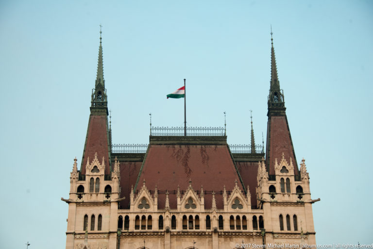 Red-tiled Parliament roof between twin spires, with the Hungarian flag flying above decorative iron cresting.