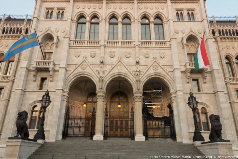 Arched Hungarian Parliament entrance with stone steps, black lion sculptures, and Hungarian and blue-and-gold flags.